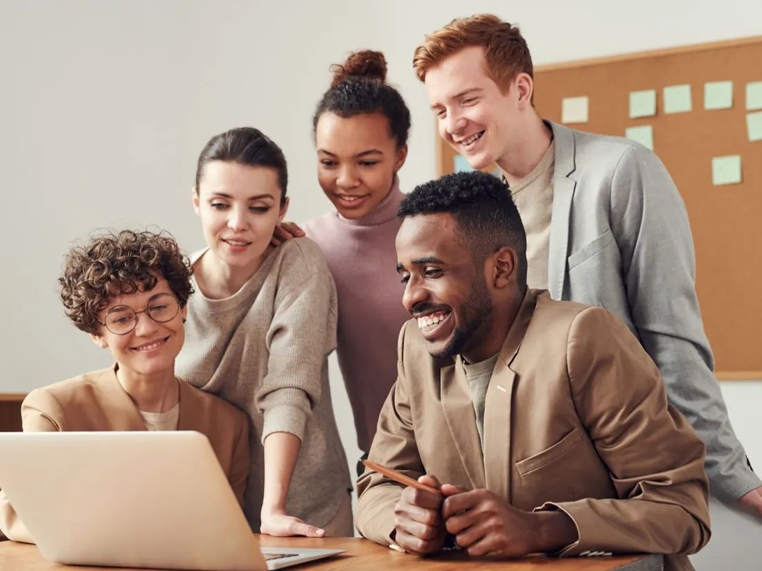 Equipe reunida e sorrindo diante de um laptop, num ambiente de trabalho acolhedor